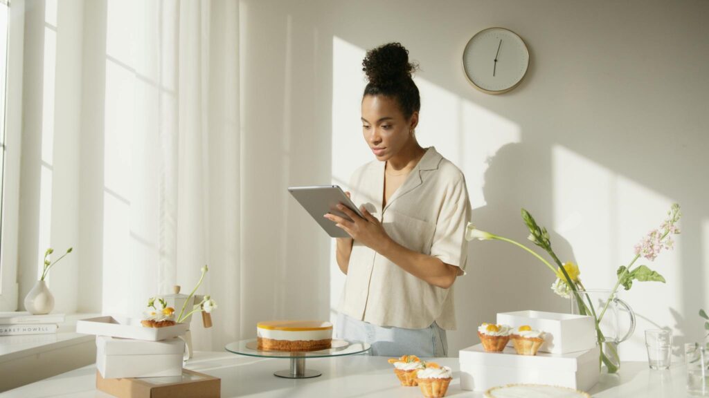 A woman using a tablet in a bright and modern kitchen setting, surrounded by cakes and flowers, depicting a lifestyle scene.